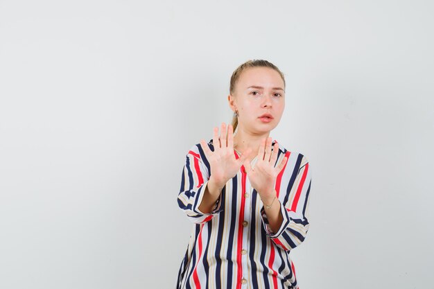 Young woman in striped blouse showing stop gestures withboth hands and looking frightened