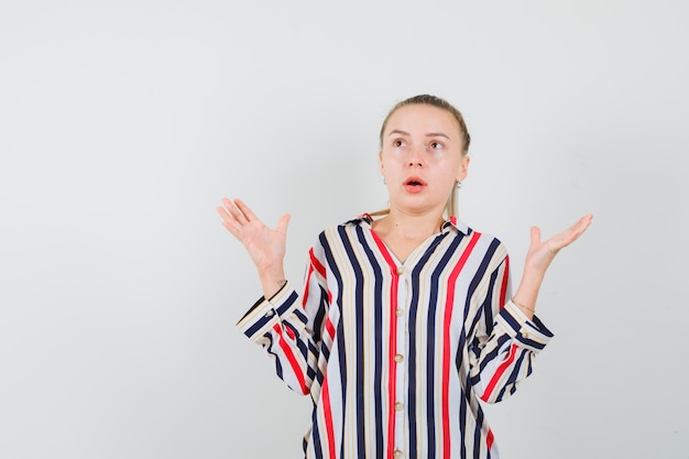 Young woman in striped blouse shrugging her shoulders and raising both hands and looking surprised