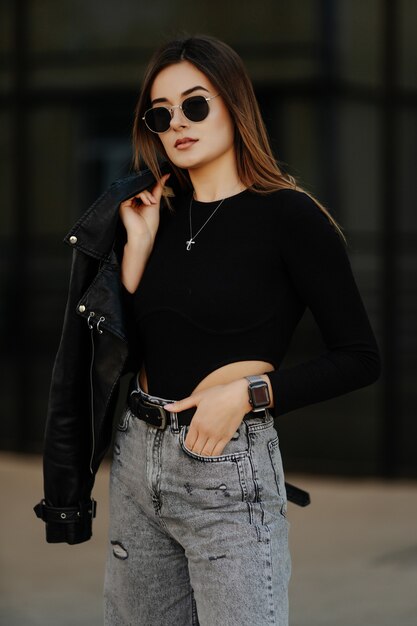 Young woman in sunglasses, hat and black leather jacket posing outdoor.