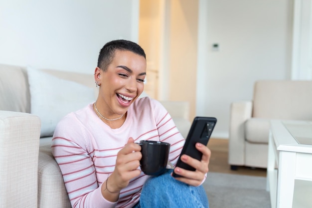 A young woman in a sweater with a cup in her hands looks at the phone while sitting on the couch