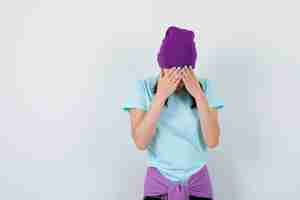 Free photo young woman in t-shirt, beanie bending head down, covering face with hands and looking depressed , front view.