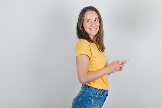 Young woman in t-shirt, shorts looking away with smartphone in hands and looking cheery .