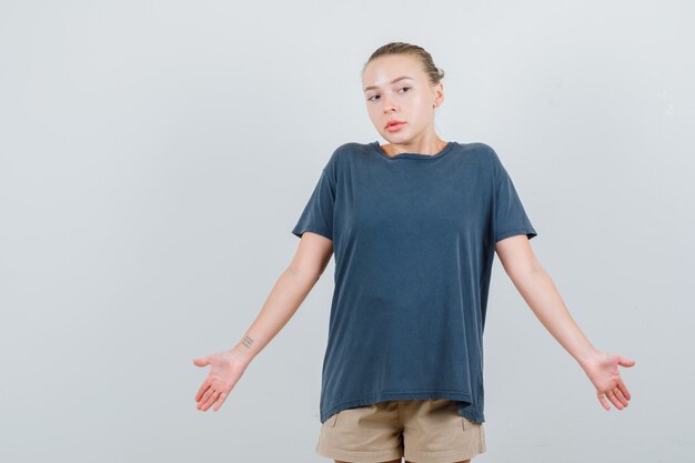 Young woman in t-shirt, shorts showing helpless gesture by shrugging and looking confused