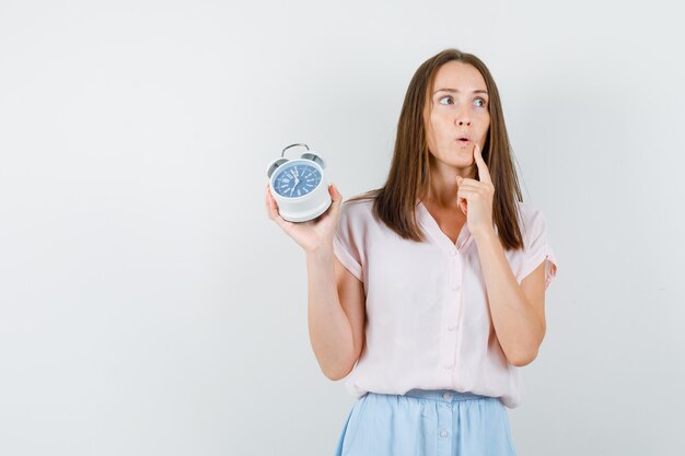 Young woman in t-shirt, skirt holding alarm clock and looking pensive , front view.