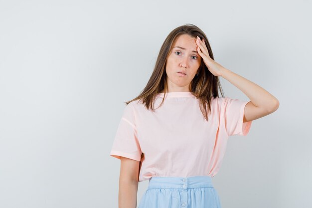 Young woman in t-shirt, skirt touching her forehead and looking puzzled , front view.