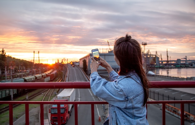 Free photo a young woman take a photo of a beautiful sunset from a bridge on her phone.