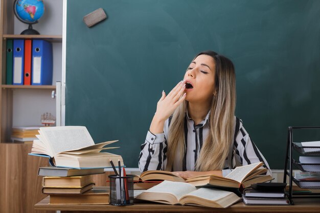 young woman teacher sitting at school desk in front of blackboard in classroom among books on her desk looking tired and overworked yawning covering mouth with hand