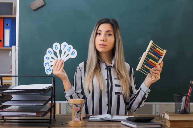 young woman teacher sitting at school desk in front of blackboard in classroom explaining lesson holding number plates and abacus looking confident