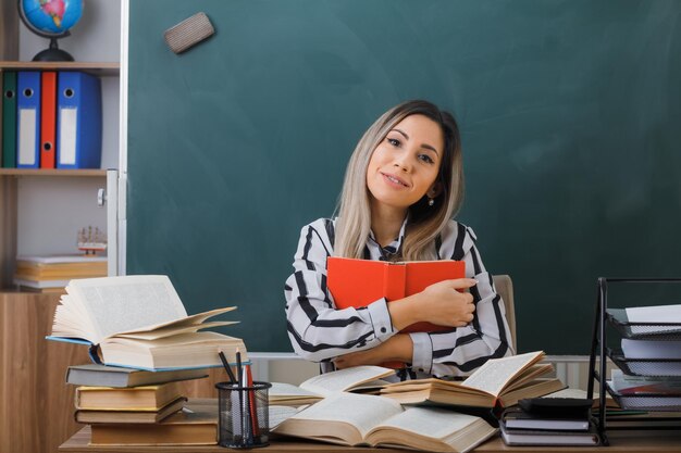 young woman teacher sitting at school desk in front of blackboard in classroom holding book looking at camera smiling friendly happy and positive