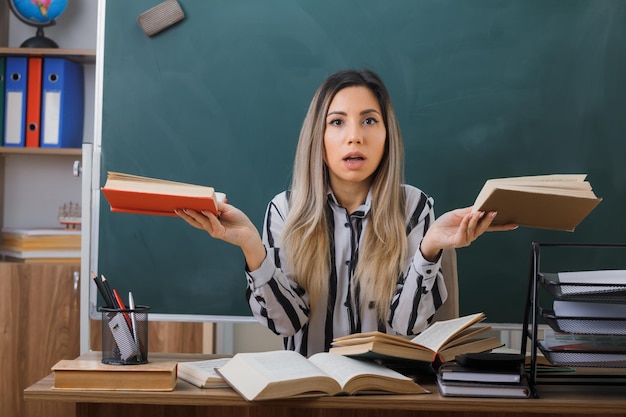 young woman teacher sitting at school desk in front of blackboard in classroom holding books looking at camera confused trying to make a choice