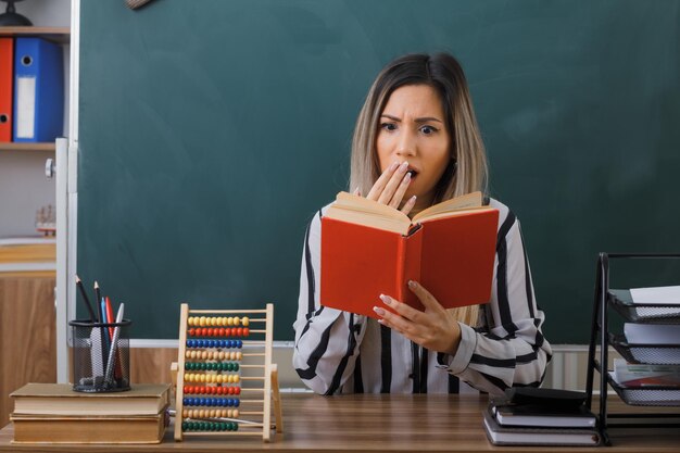 young woman teacher sitting at school desk in front of blackboard in classroom reading book preparing for lesson looking worried and shocked