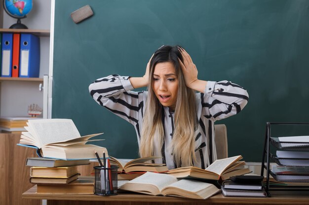 young woman teacher sitting at school desk in front of blackboard in classroom with books looking at mess on her desk being confused and worried with hands on her head