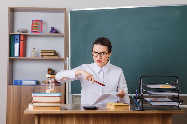 Young woman teacher wearing glasses checking homework of students being displeased and angry sitting at school desk in front of blackboard in classroom