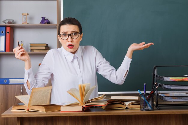 Young woman teacher wearing glasses checking homework of students looking confused and displeased sitting at school desk in front of blackboard in classroom