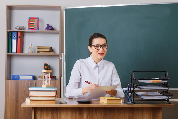 Young woman teacher wearing glasses checking homework of students lookingat camera smiling pleased and happy sitting at school desk in front of blackboard in classroom