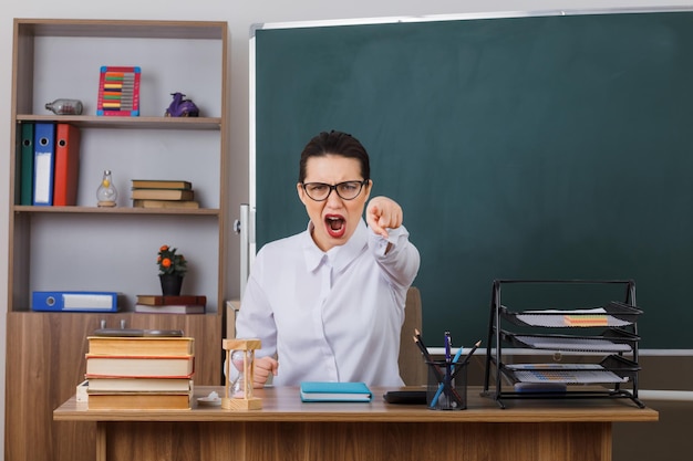 Young woman teacher wearing glasses explaining lesson looking angry and frustrated shouting pointing with index finger at camera sitting at school desk in front of blackboard in classroom