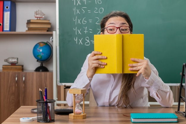 Young woman teacher wearing glasses holding book preparing for lesson being confused and worried sitting at school desk in front of blackboard in classroom