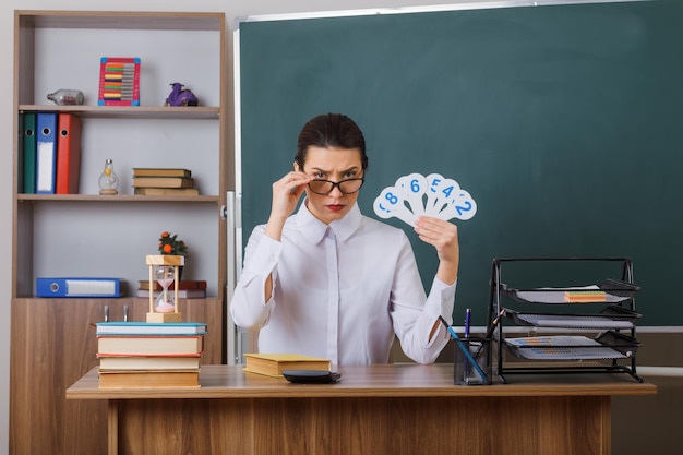 Young woman teacher wearing glasses holding number plates explaining lesson looking frowningly sitting at school desk in front of blackboard in classroom