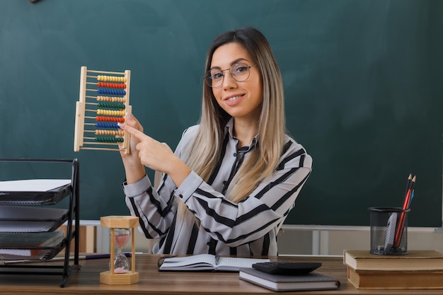 Free photo young woman teacher wearing glasses sitting at school desk in front of blackboard in classroom explaining lesson holding abacus smiling confident