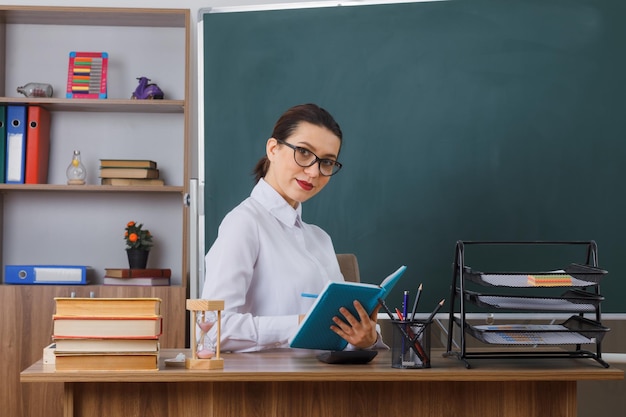 Young woman teacher wearing glasses sitting at school desk in front of blackboard in classroom explaining lesson looking confident