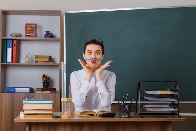 Free photo young woman teacher wearing glasses sitting at school desk with book in front of blackboard in classroom holding pen between lips and nose having fun crossing eyes