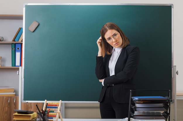 Free Photo young woman teacher wearing glasses standing near blackboard in classroom explaining lesson looking confused scratching her head trying to remember important thing