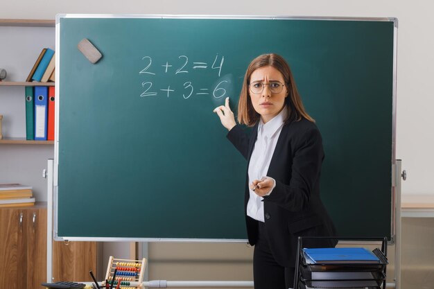 Young woman teacher wearing glasses standing near blackboard in classroom explaining lesson pointing at blackboard with pointer looking disappointed