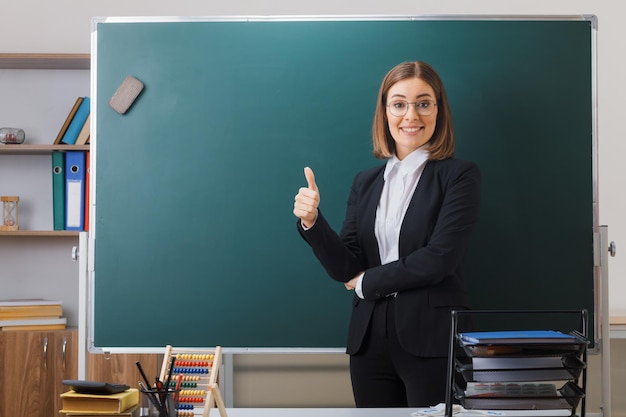 Young woman teacher wearing glasses standing near blackboard in classroom explaining lesson showing thumb up smiling cheerfully happy and positive