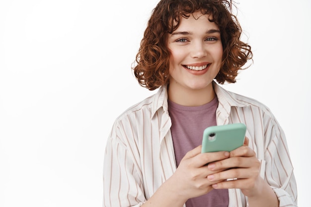 Free Photo young woman texting on mobile phone. girl smiles at camera, uses smartphone, reading message or using application, order taxi app, standing over white background