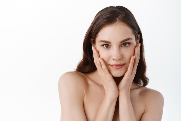 Young woman touching moisturized clean and hydrated facial skin holding hands on soft cheeks gazing at camera standing with naked shoulders against white background