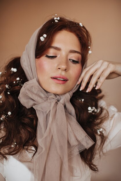 Young woman in transparent scarf on her head gently touches her face and modestly looks down.