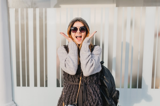 Free photo young woman in trendy sunglasses enjoying good weather during morning walk. outdoor portrait of excited woman in gray sweater carrying black backpack and posing with surprised face expression.