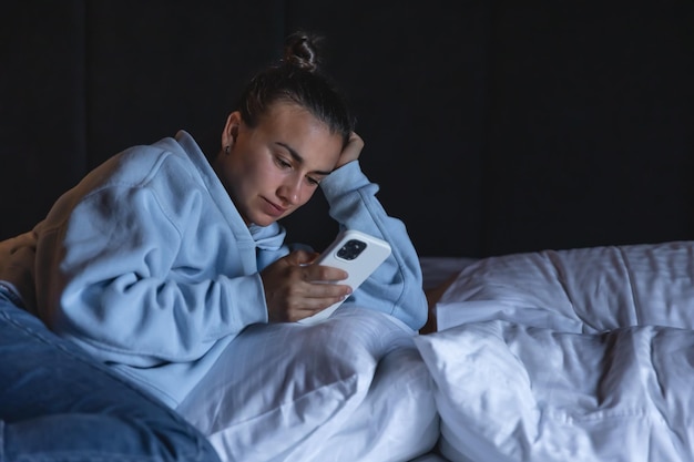 A young woman uses a smartphone while lying in bed in the evening