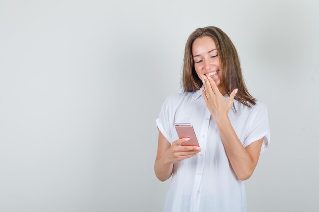 Young woman using smartphone with hand on mouth in white t-shirt and looking happy