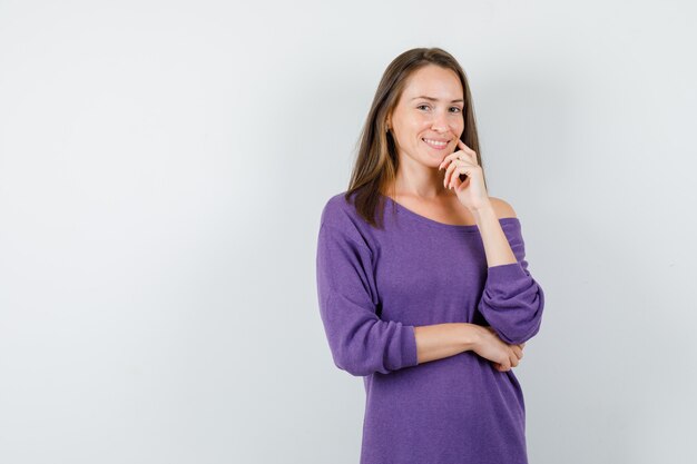 Young woman in violet shirt standing in thinking pose and looking cheerful , front view.