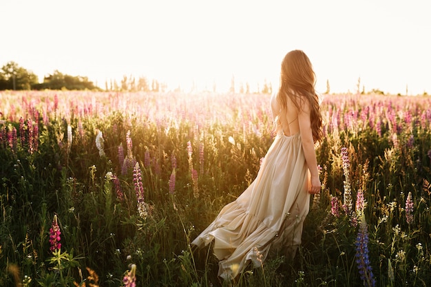 Young woman walking on flower field at sunset on background.