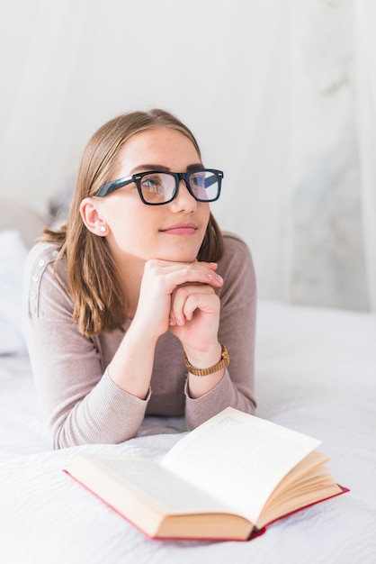 Free photo young woman wearing black eyeglasses daydreaming while reading book on bed