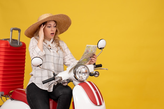 Young woman wearing hat and sitting on motorcycle and looking at map with confused facial epression on yellow 