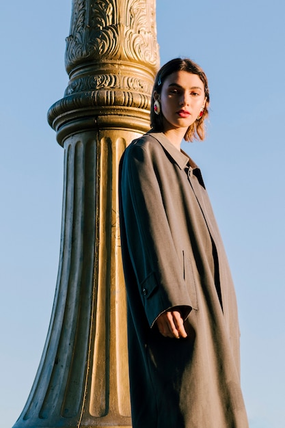 Young woman wearing long coat standing near the pillar against blue sky