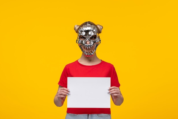 Young woman wearing terrifying golden mask and holding piece of paper yellow background horror