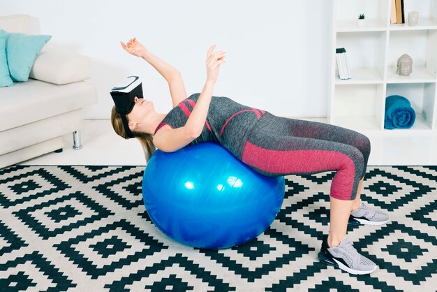 Young woman wearing virtual reality glasses stretching over pilates ball touching the hands in the air