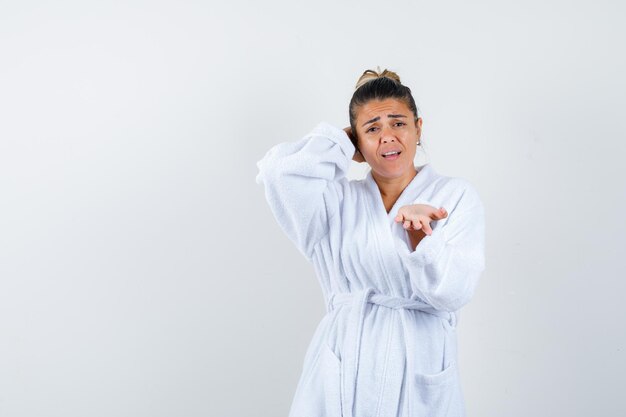 Young woman in white bathrobe stretching hand as receiving something while holding hand behind head and looking annoyed