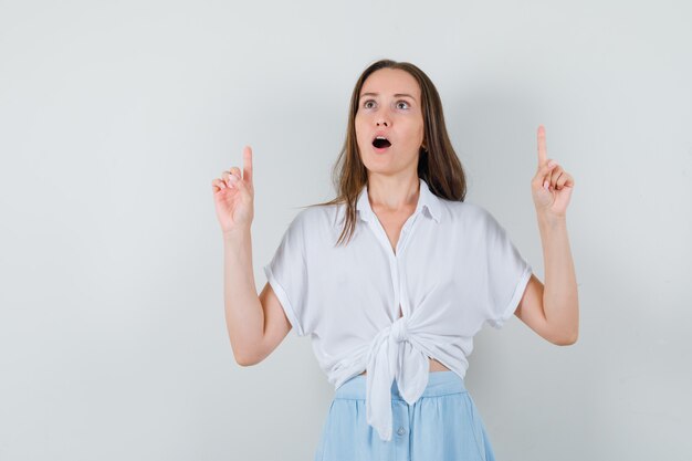 Young woman in white blouse and light blue skirt pointing up with index fingers and looking surprised