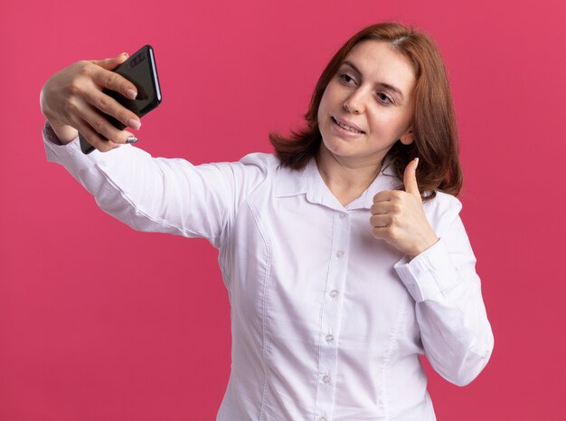 Young woman in white shirt holding smartphone doing selfie smiling showing thumbs up standing over pink wall