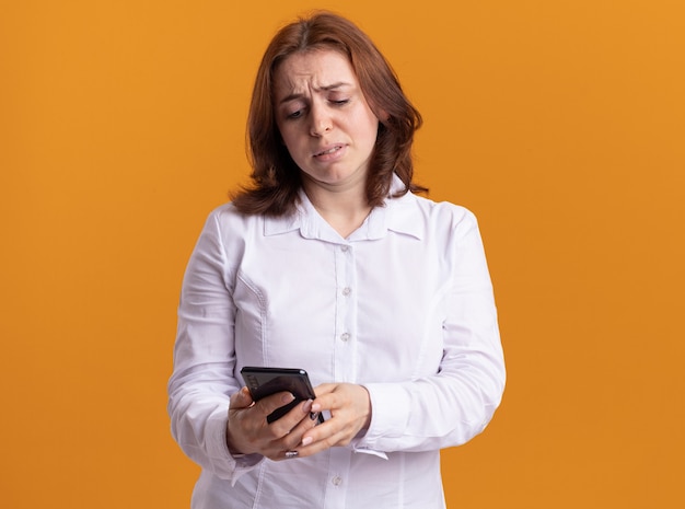 Young woman in white shirt holding smartphone looking at it with sad expression standing over orange wall