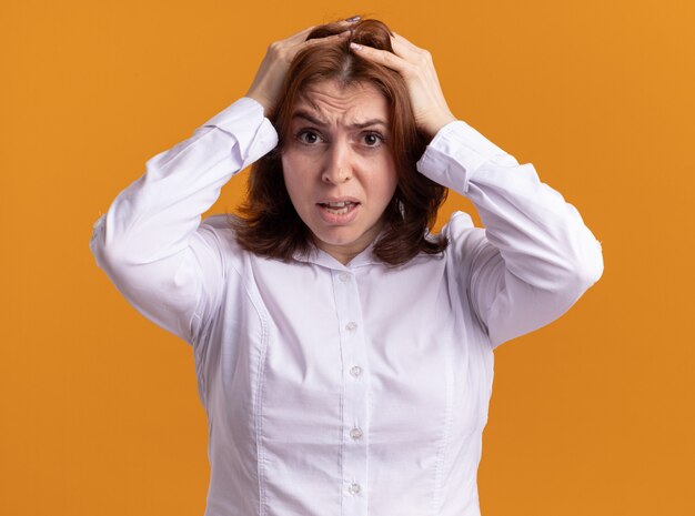 Young woman in white shirt lookign at front confused and frustrated with hands on her head for mistake standing over orange wall