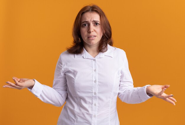 Young woman in white shirt looking at front confused spreading arms to the sides standing over orange wall