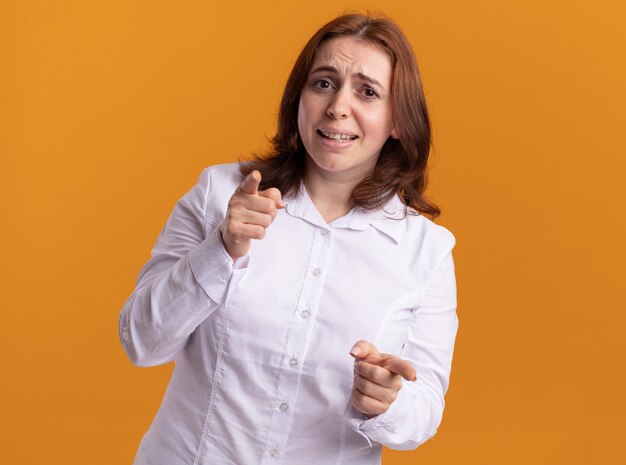 Young woman in white shirt looking at front smiling pointing with index fingers at front standing over orange wall