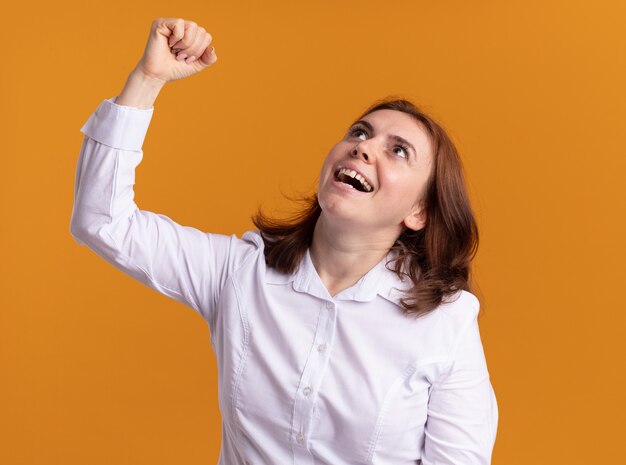 Young woman in white shirt looking up happy and excited raising fist like a winner standing over orange wall