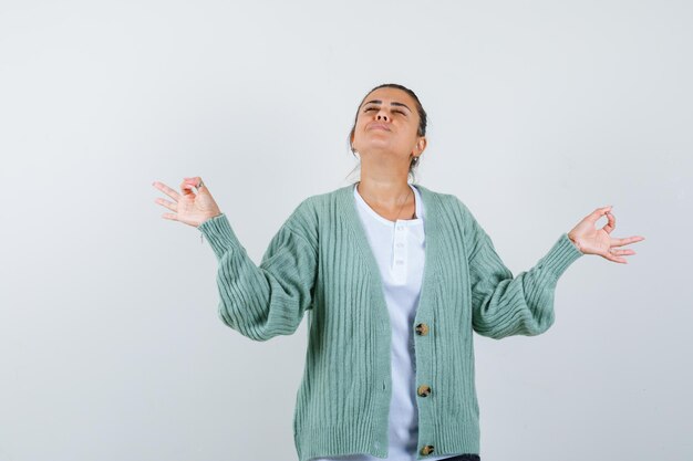 Young woman in white shirt and mint green cardigan standing in meditating pose and looking calm